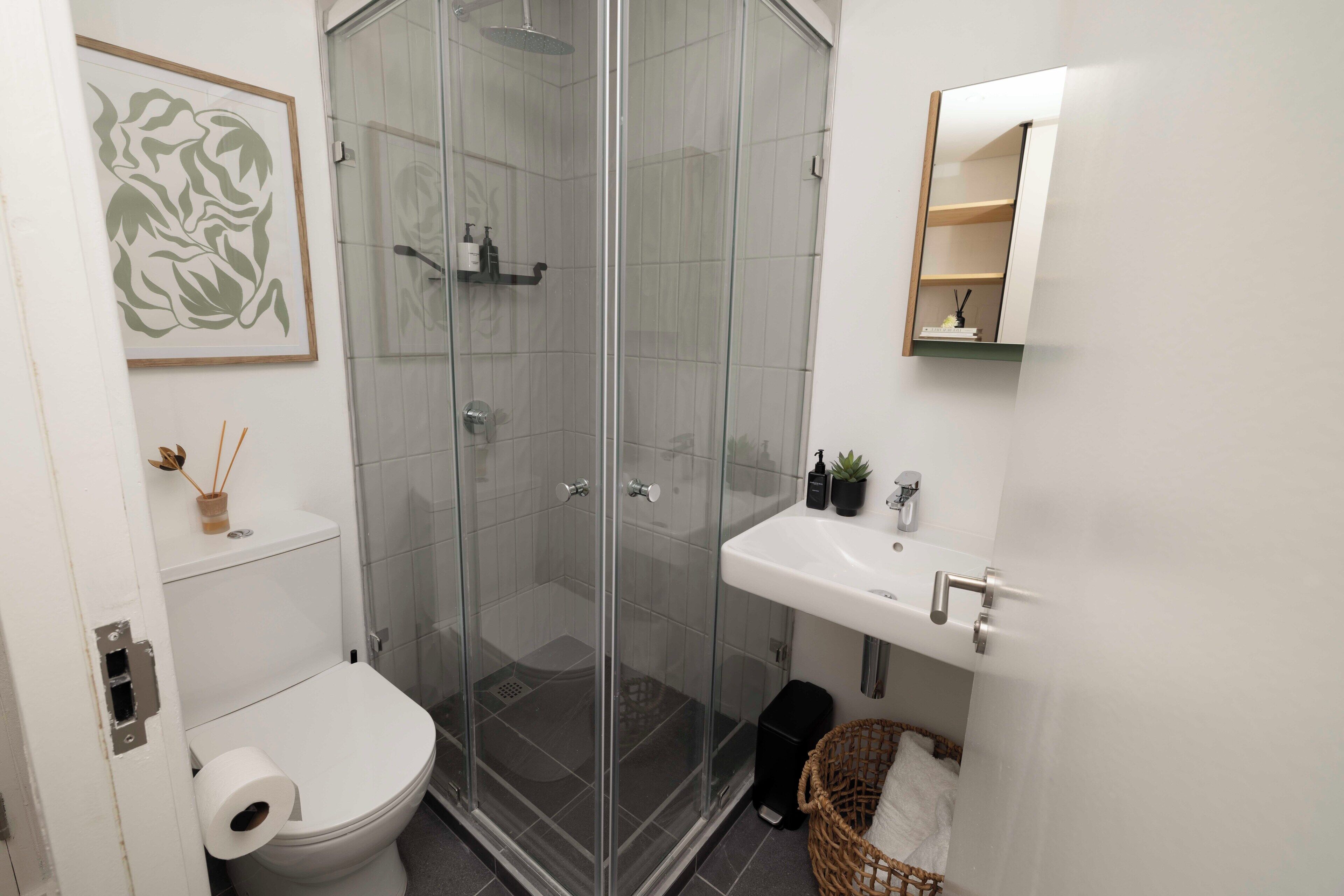 A modern bathroom with a glass-enclosed shower, a white toilet, a sink with a mirror, and a small shelf with toiletries.