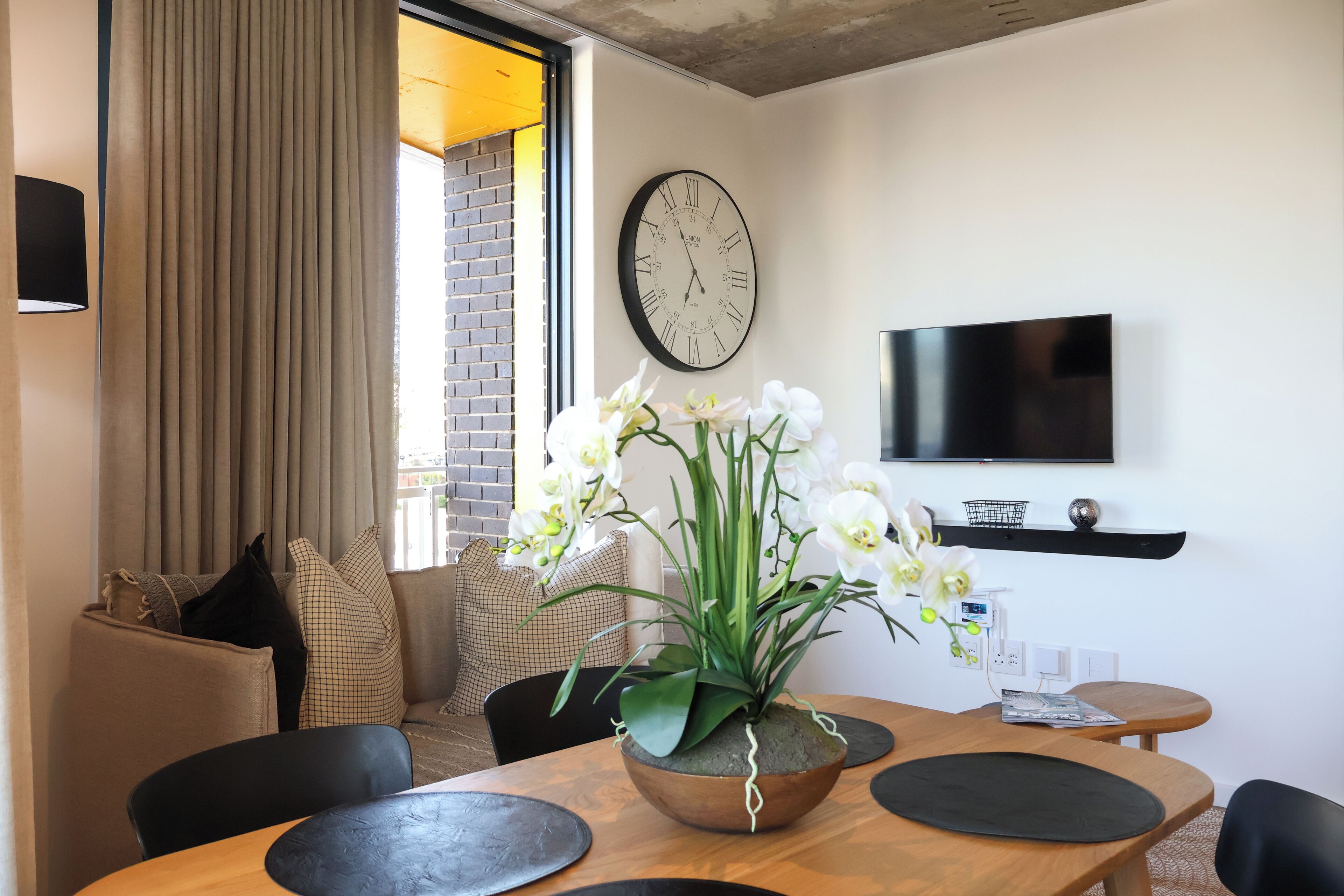 A modern living room with a wooden dining table, a large wall clock, and a flat-screen TV mounted on the wall.