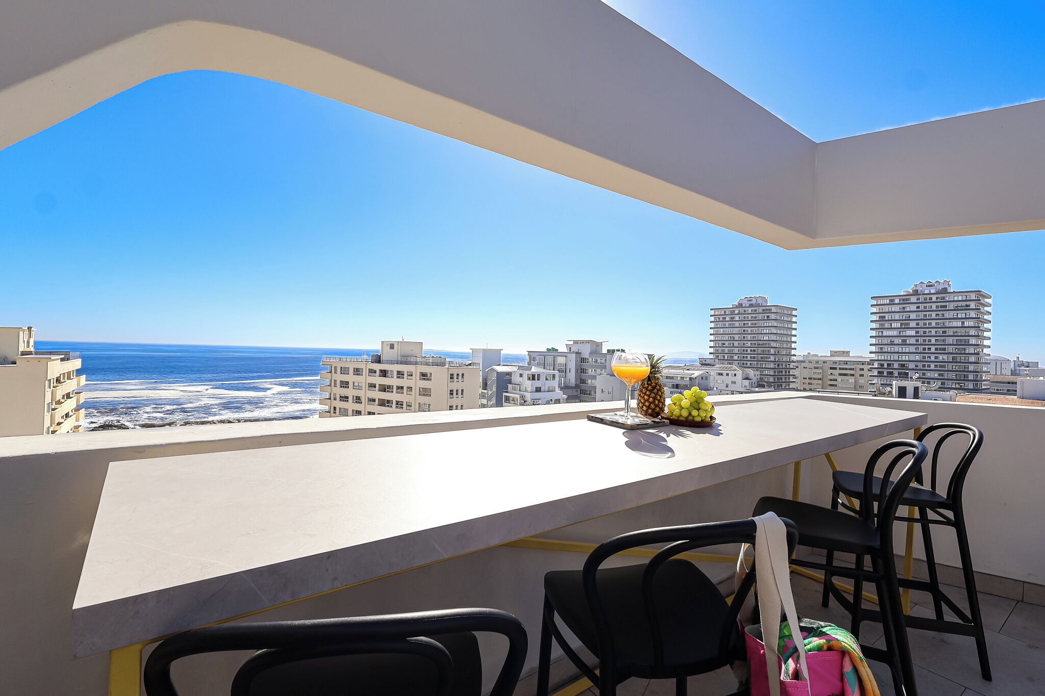 Balcón con vista al océano y al paisaje urbano, con una mesa, sillas y un frutero.
