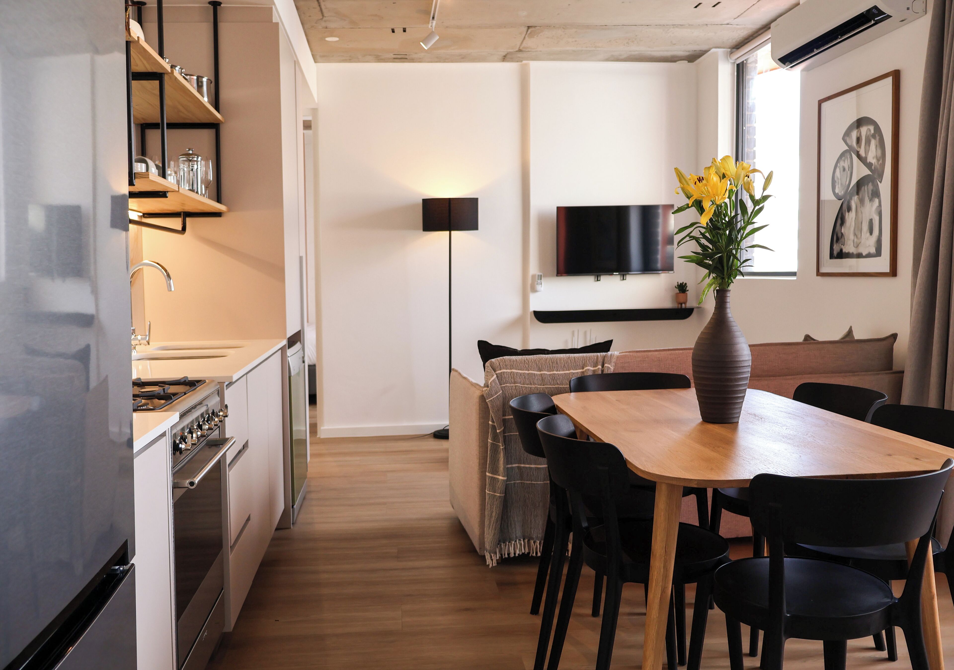 A modern kitchen with built-in appliances, a dining area with a wooden table and black chairs, and a living space with a sofa and a floor lamp.