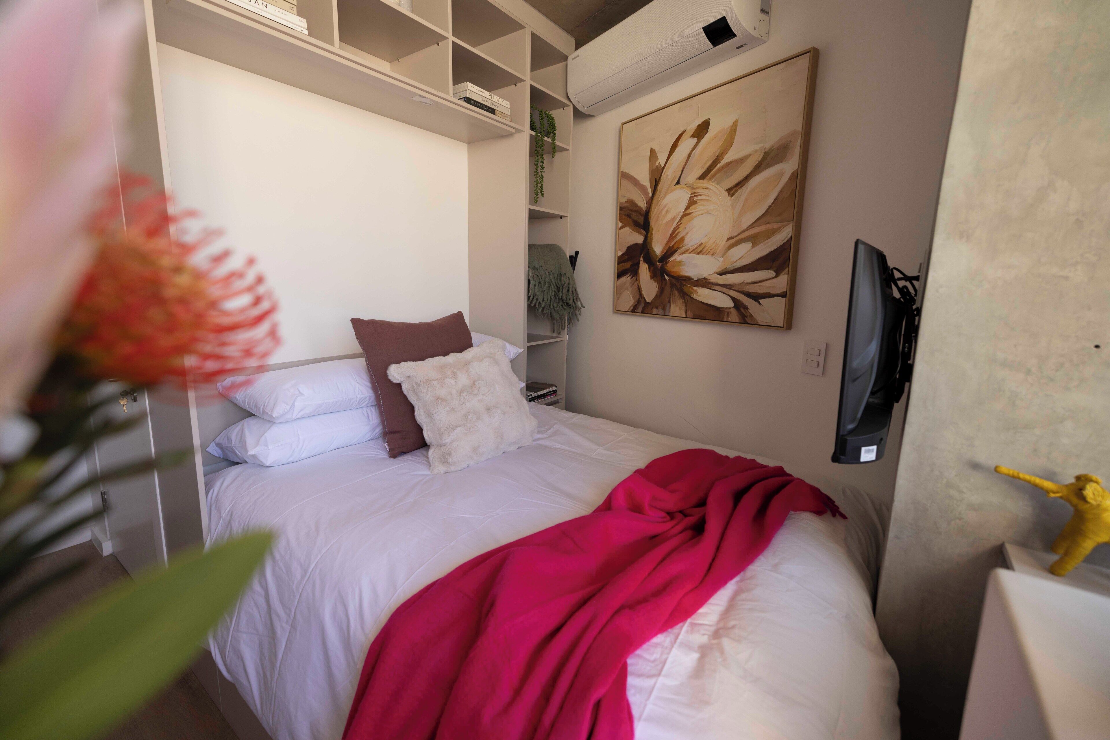 A bedroom with a bed, a red blanket, a white pillow, a brown pillow, a painting, and a television.