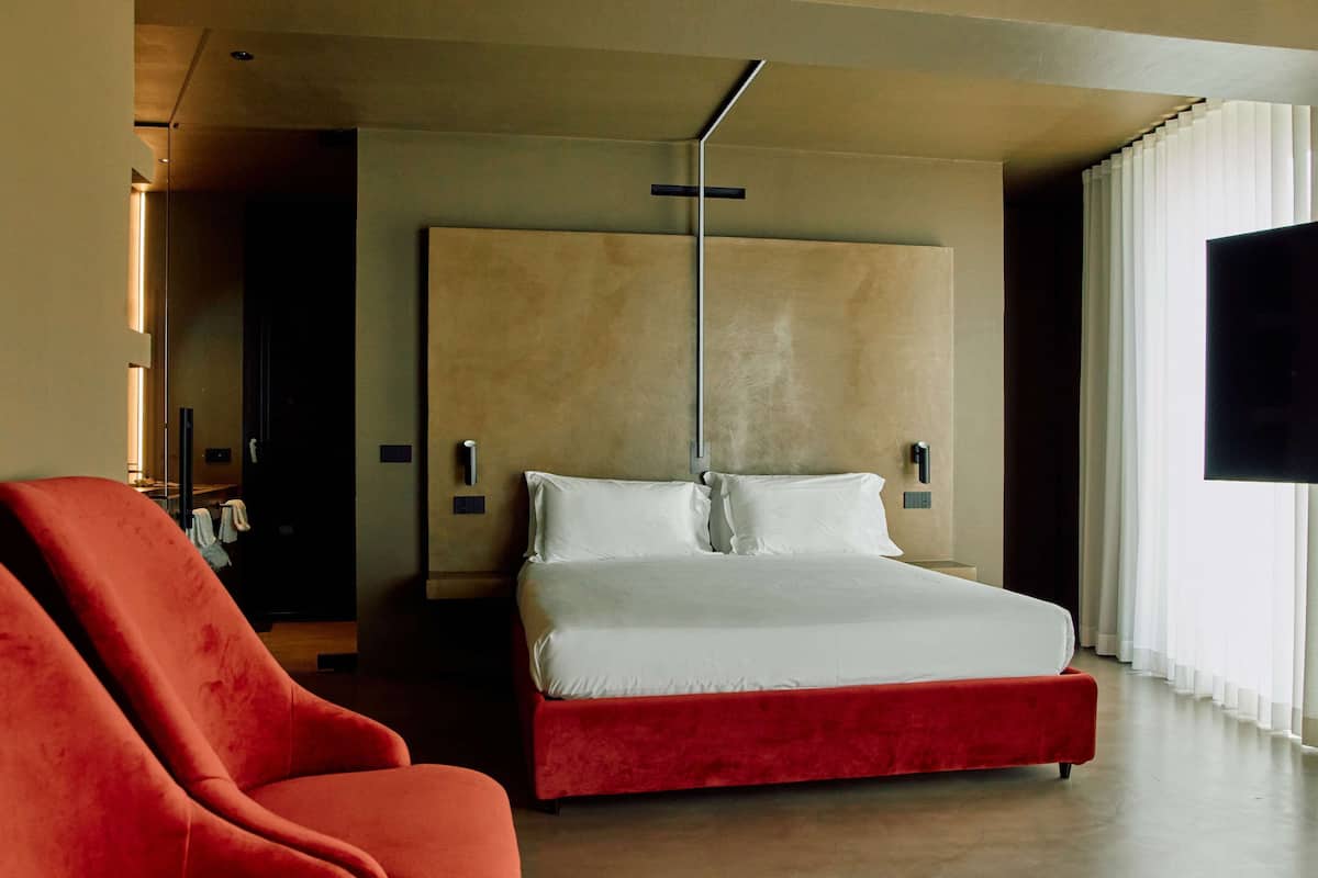 A modern hotel room with a large bed, a red chair, a flat-screen TV, and a large window with curtains.