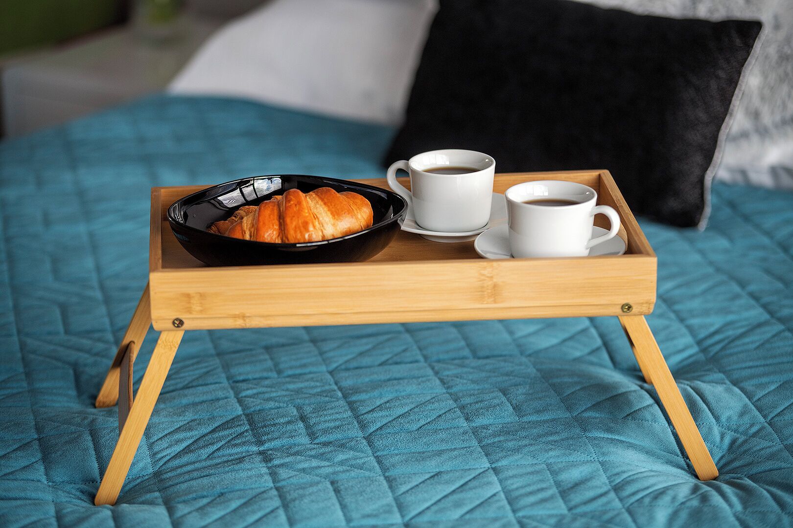 Una bandeja con croissants, dos tazas de café y un bol negro sobre una cama con colcha azul.