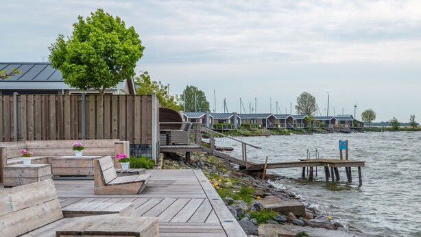 Una terraza de madera con bancos y plantas en macetas, con vista a un cuerpo de agua y un muelle que se extiende hacia el agua.