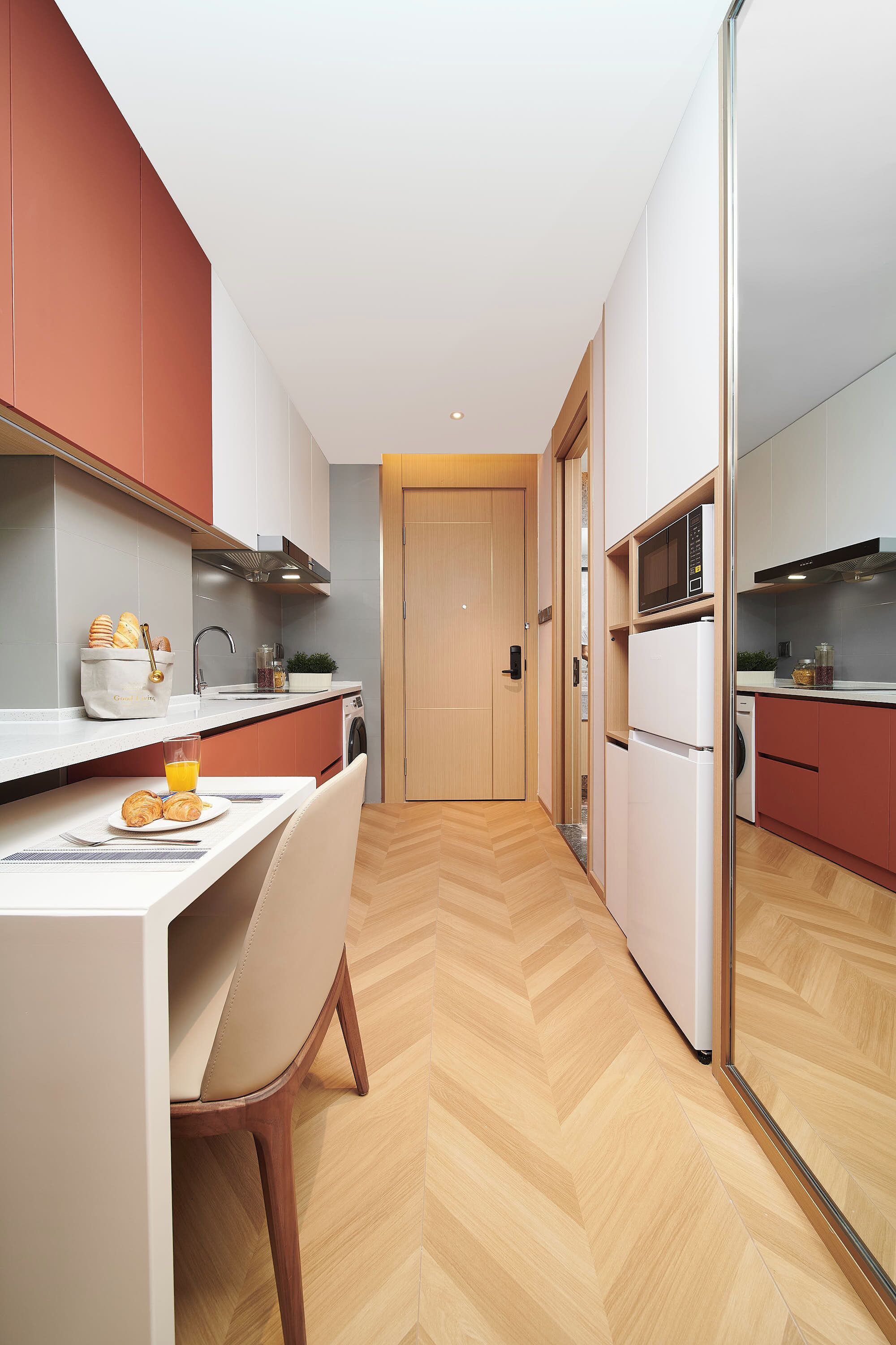 A modern kitchen with a microwave, refrigerator, and sink. There is a dining table with a chair and a glass of orange juice. The floor has a herringbone pattern.