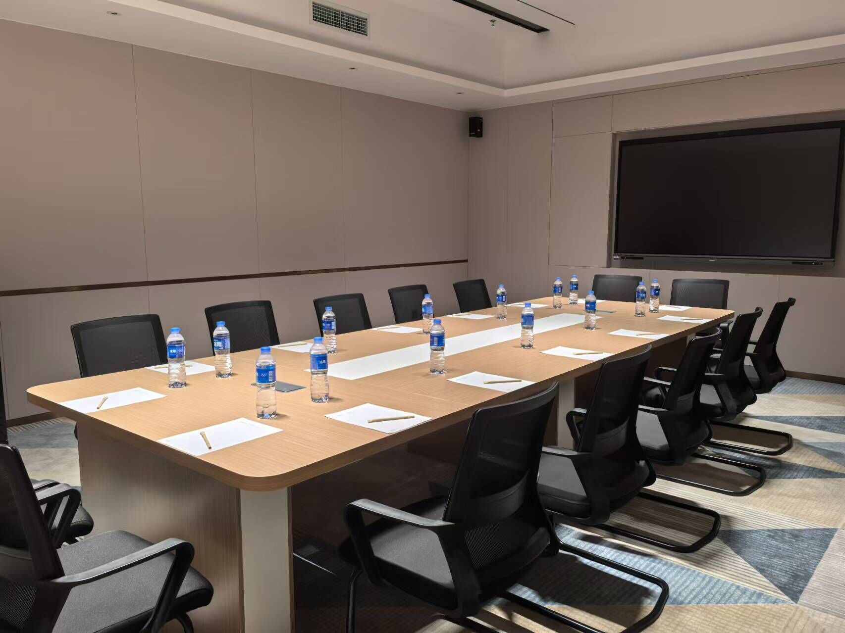 Un salón de conferencias con una larga mesa, sillas negras, botellas de agua y una pantalla para proyector.
