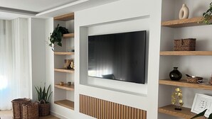 Ett modernt vardagsrum med en stor platt-TV som är monterad på en vit vägg, trähyllor med dekorativa föremål, ett träbord och en krukväxt.