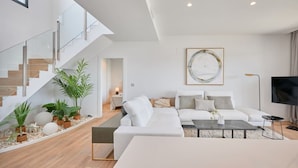 En moderne stue med en hvid sofa, et sofabord og et fjernsyn. Der er en trappe med glasgelændere og en plante på repos.'
