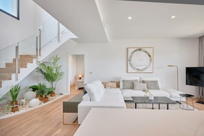 Una sala moderna con un sofá blanco, una mesa de centro y un televisor. Se observa una escalera con barandillas de cristal y una planta en el rellano.