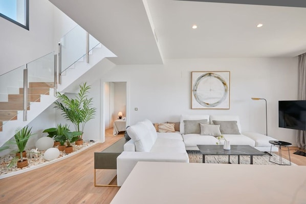 A modern living room with a white sofa, a coffee table, and a TV. There is a staircase with glass railings and a plant on the landing.