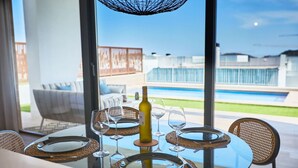 A dining table set for four with wine glasses and a bottle, overlooking a swimming pool and outdoor seating area. - Campana Bay Villa (Finestrat)