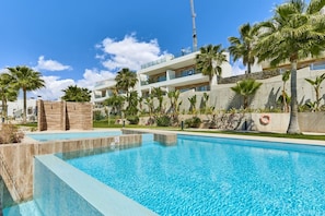 Una piscina con superficie de agua azul cristalina, rodeada de palmeras y un edificio moderno con balcones.