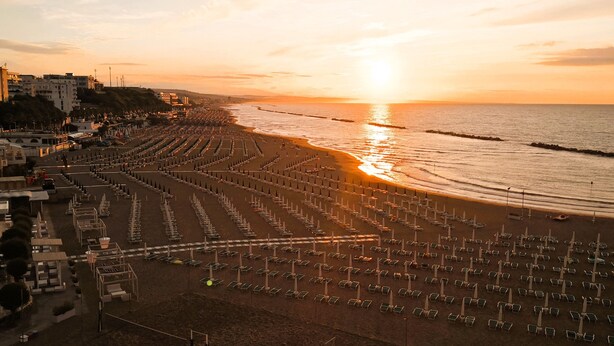 Una spiaggia con file di lettini e ombrelloni, edifici sullo sfondo e un tramonto sull'oceano.