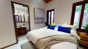 Une chambre à coucher avec un lit, une table de chevet, une fenêtre, un miroir et une salle de bain visible à travers une porte ouverte.