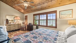 Een slaapkamer met een houten plafond, een groot bed met een geruite quilt, een dressoir, een stoel en een raam met uitzicht op de omgeving.