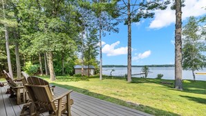 Een terras met Adirondack-stoelen met uitzicht op een meer, omringd door bomen en een klein huisje.