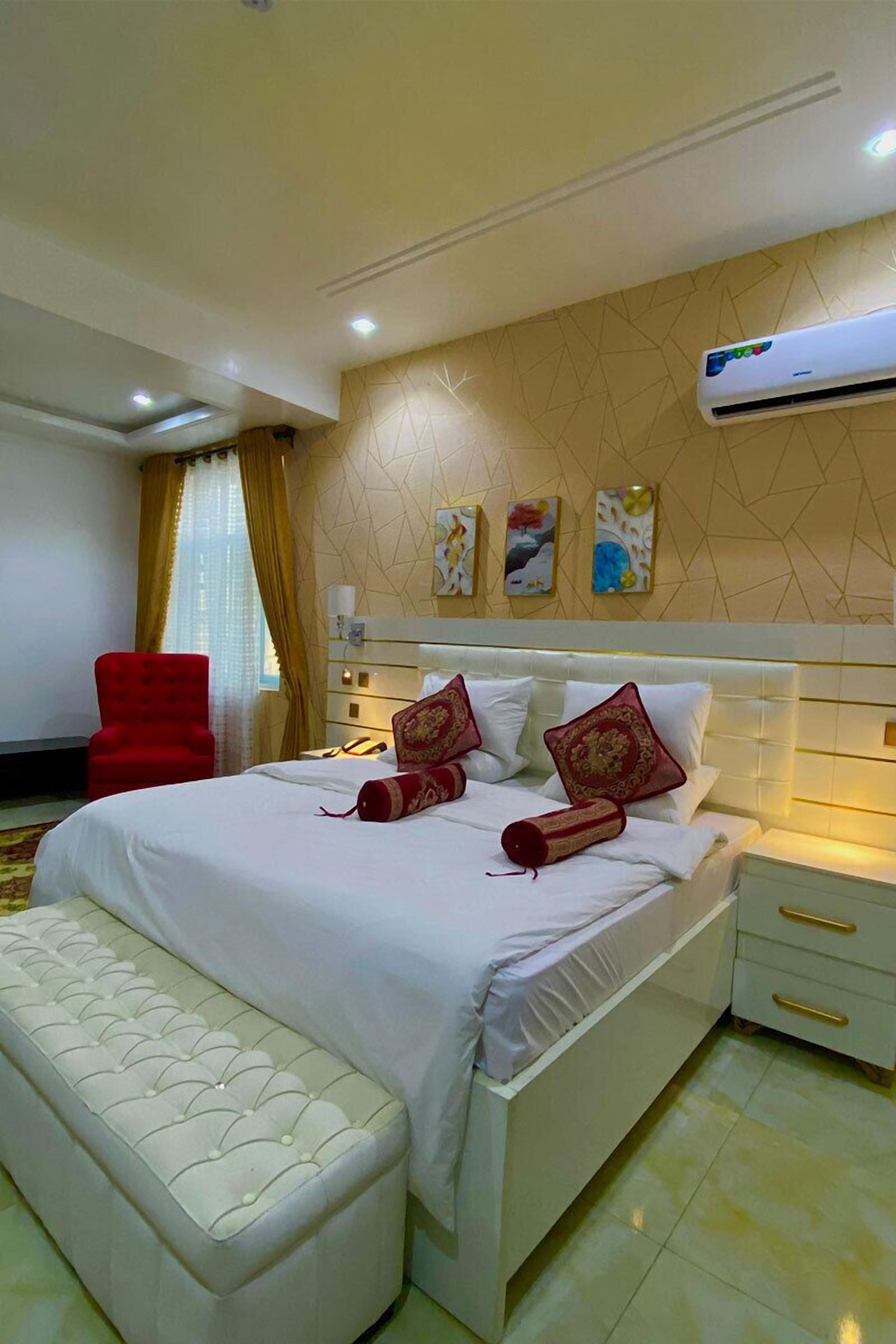 A hotel room with a bed, a red armchair, a bedside table, and an air conditioning unit.