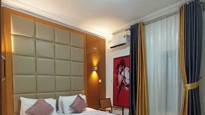 A hotel room with a large bed, a headboard, a nightstand, a chair, a wall-mounted air conditioner, and a window with curtains. - AJ'S Hotel & Suites (Abuja)