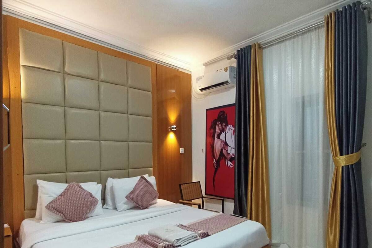 Une chambre d’hôtel avec un grand lit, une tête de lit, une table de chevet, une chaise, un climatiseur fixé au mur et une fenêtre avec des rideaux.