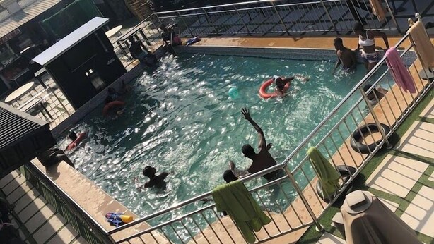 A swimming pool area with multiple people swimming and playing, a covered seating area, and a view of the surrounding landscape. - AJ'S Hotel & Suites (Abuja)