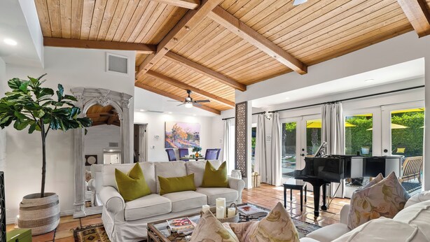 A spacious living area with a white sofa, green cushions, a coffee table, and a large plant. A piano is visible in the background, and there is a view of greenery outside. - Good Vibes Only – Historic Spanish Hacienda with Pool, Spa, Fire Pit & Mountain Views (Palm Springs)