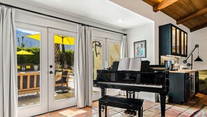 A spacious room with a grand piano, a dining area with a table and chairs, and a view through glass doors to an outdoor patio with umbrellas. - Good Vibes Only – Historic Spanish Hacienda with Pool, Spa, Fire Pit & Mountain Views (Palm Springs)
