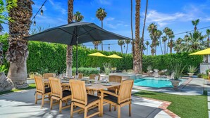 A poolside patio with a wooden table and chairs, surrounded by palm trees and a swimming pool. - Good Vibes Only – Historic Spanish Hacienda with Pool, Spa, Fire Pit & Mountain Views (Palm Springs)