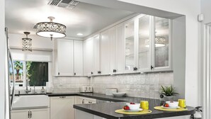 A modern kitchen with white cabinets, a black countertop, and two bar stools. - Good Vibes Only – Historic Spanish Hacienda with Pool, Spa, Fire Pit & Mountain Views (Palm Springs)