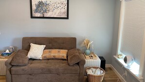 A cozy living room with a brown sofa, a framed artwork above it, a side table with a vase and a basket, and a window with blinds.