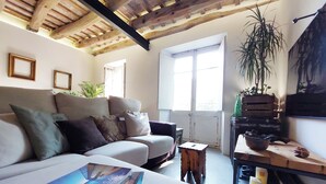 A cozy living room with a sofa, a coffee table with books, a wooden side table, and a large window with natural light.