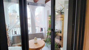 An open-plan living space with a wooden dining table, a white rug, and a glass door leading to an outdoor area with plants and a view of buildings.