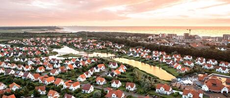 Ett flygbildsbild av ett bostadsområde med hus, en vattenyta och en avlägsen stadsbild i solnedgången.
