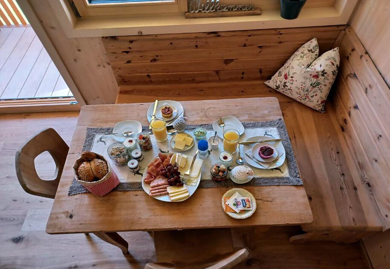 A wooden dining table with a breakfast spread including bread, cheese, meats, and fruits.