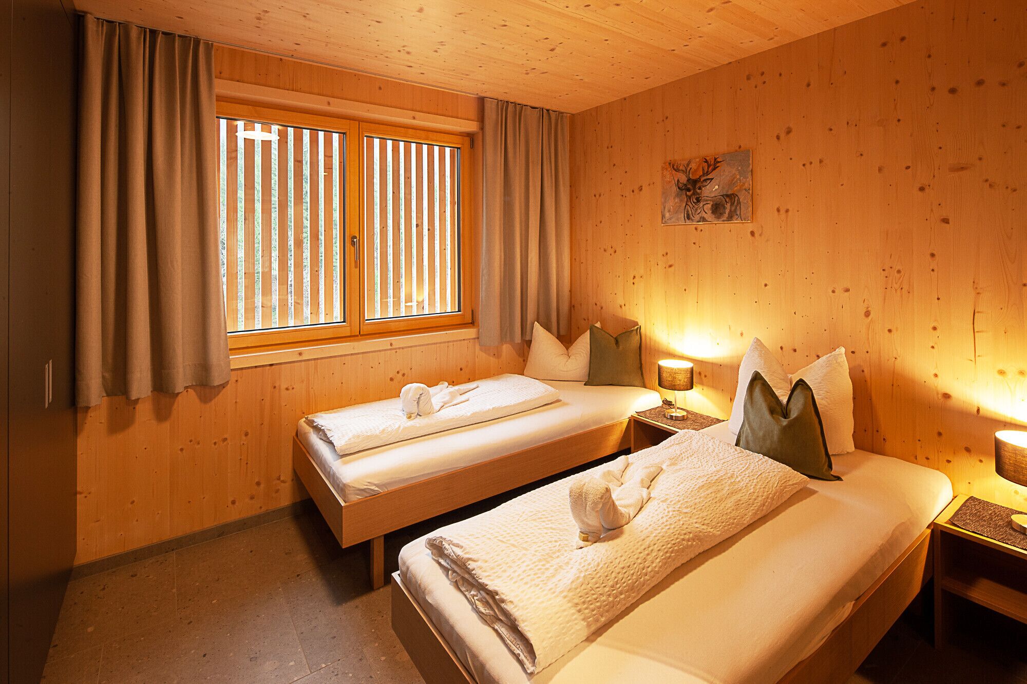 A wooden cabin room with two beds, a window with curtains, and a painting on the wall.