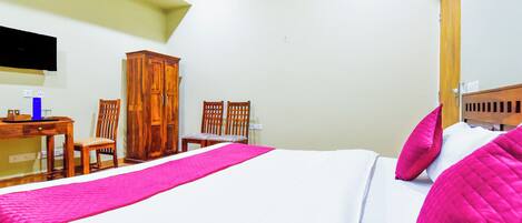 A bedroom with a bed, two chairs, a wooden wardrobe, a desk, and a flat-screen TV mounted on the wall.