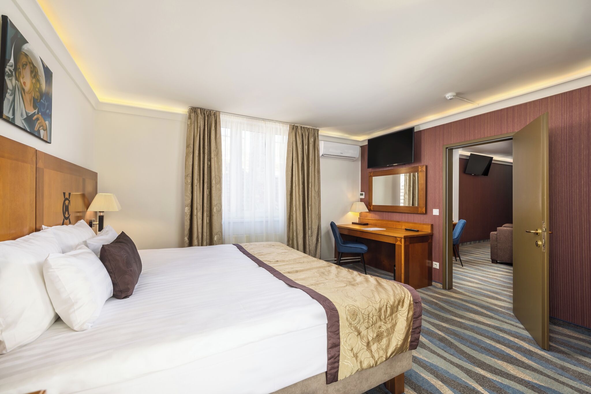 A hotel room with a large bed, a desk, a chair, a television, and an air conditioning unit.