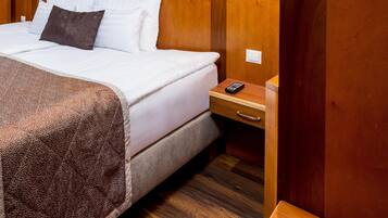 Cama bem arrumada com roupas de cama brancas e castanhas, cabeceira de madeira, mesa de cabeceira com controle remoto e luminária fixada na parede.