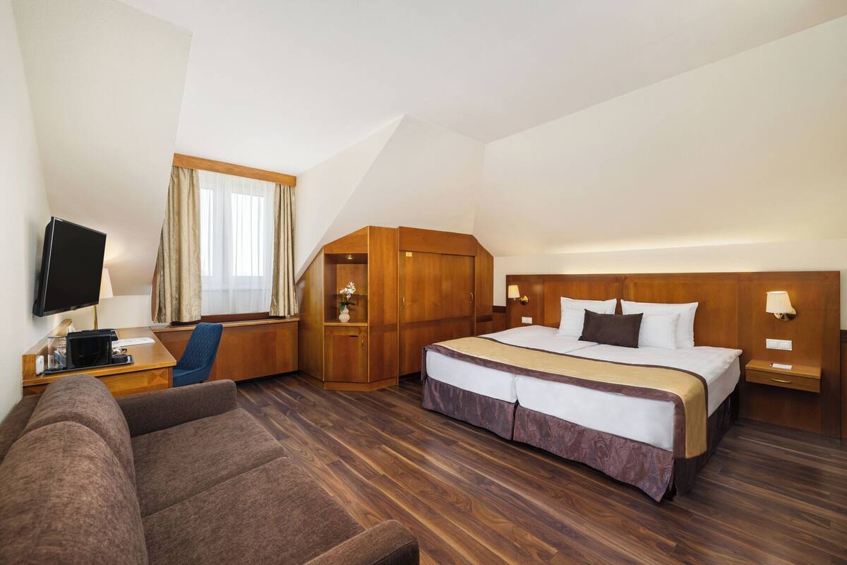 Ein Hotelzimmer mit einem großen Bett, einem Schreibtisch mit Fernseher, einem Sessel und einem Kleiderschrank.