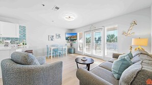 A spacious living area with a sofa, a coffee table, and a dining table with chairs. There is a television mounted on the wall, and a window with blinds.
