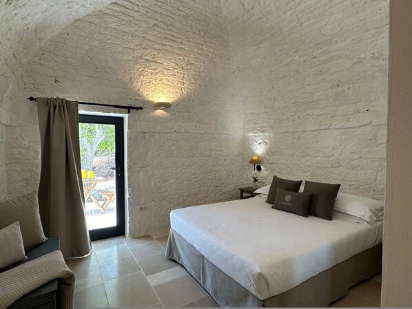 A bedroom with a bed, a nightstand, a lamp, a window with curtains, and a view of the outdoors.