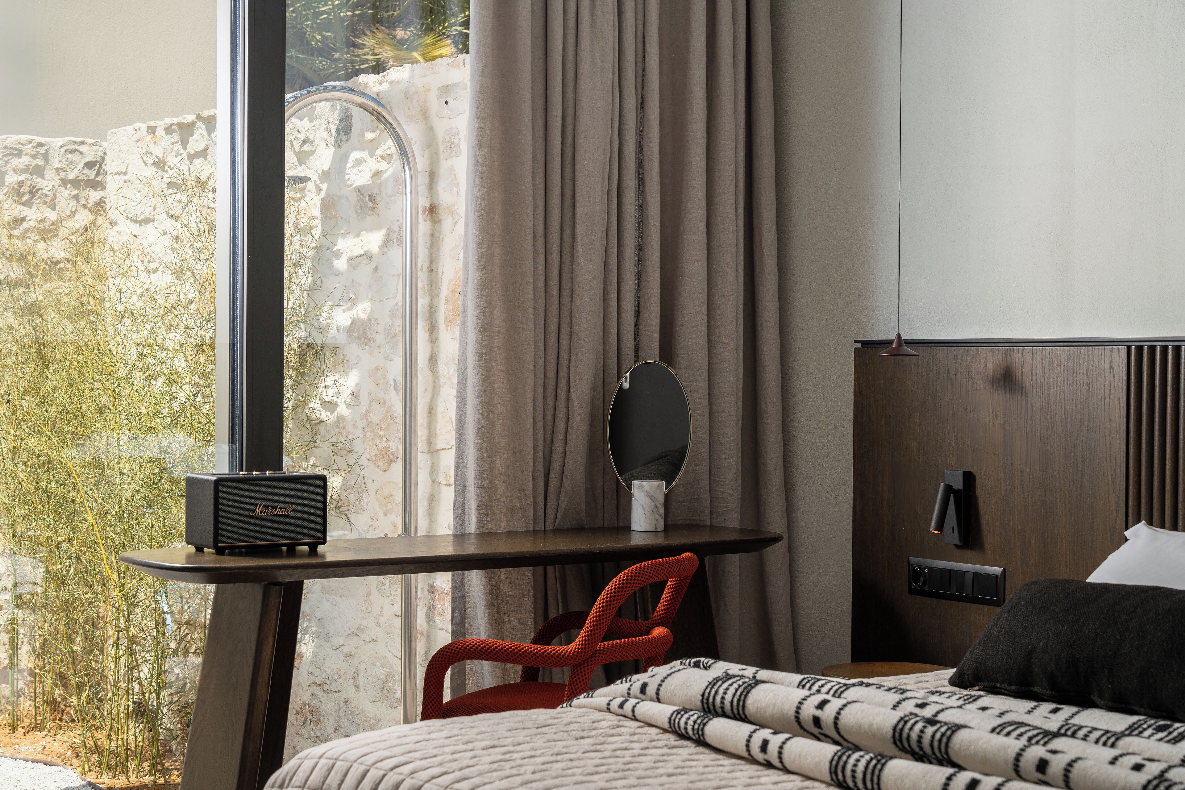 A hotel room with a bed, a desk with a chair, a small table with a radio, a mirror, and a nightstand with a lamp.