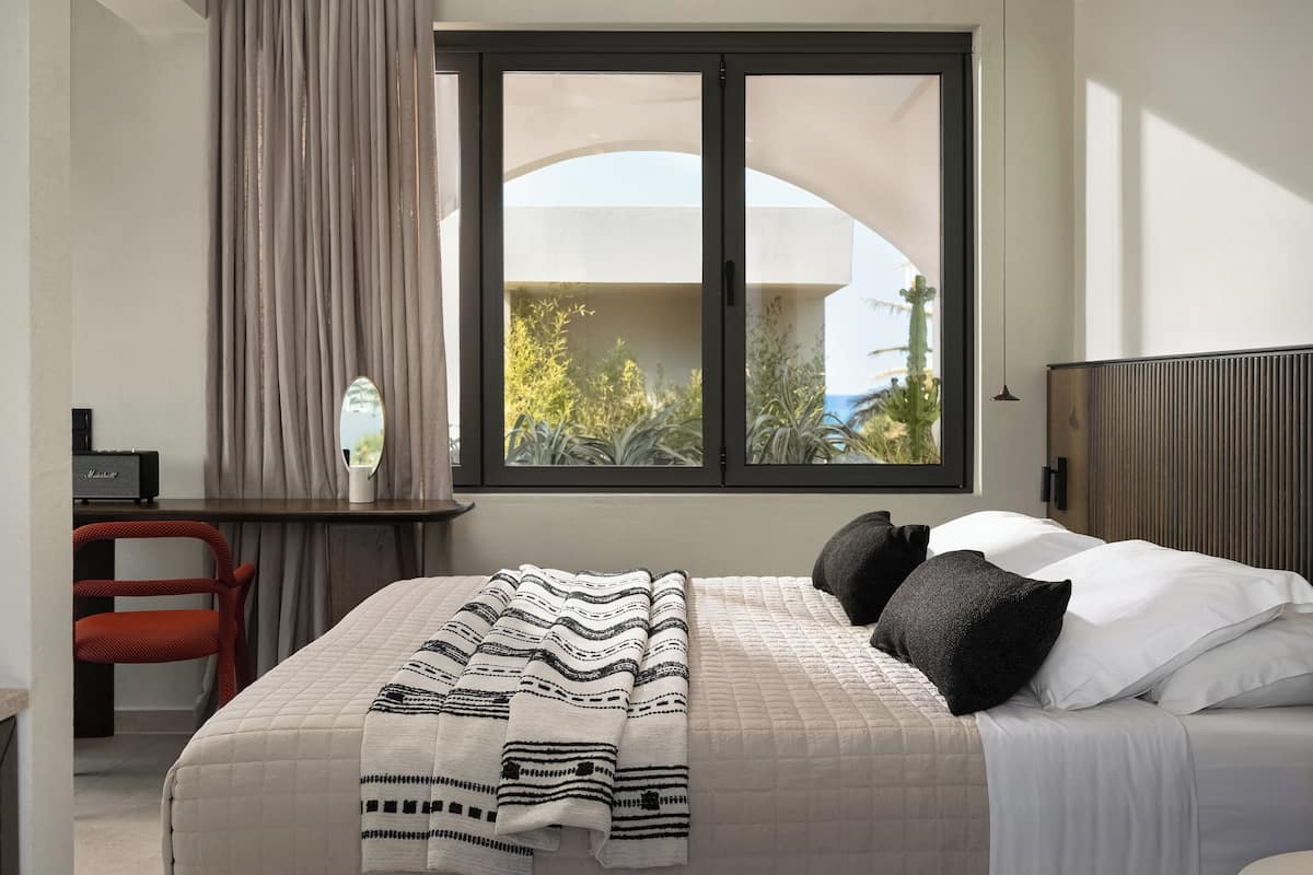A neatly made bed with a patterned blanket, a bedside table with a lamp, and a window with a view of greenery and a building.