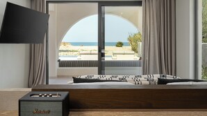 A bedroom with a large window, a flat-screen TV mounted on the wall, and a Marshall speaker on the floor. - Dyo Beach Suites (Rethymno)