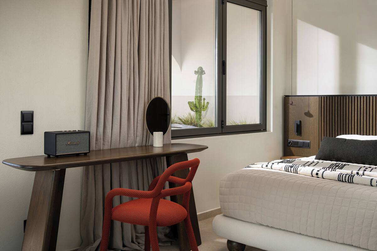 A hotel room with a bed, a desk with a radio, a chair, a window with a view of a cactus, and a mirror on the wall.