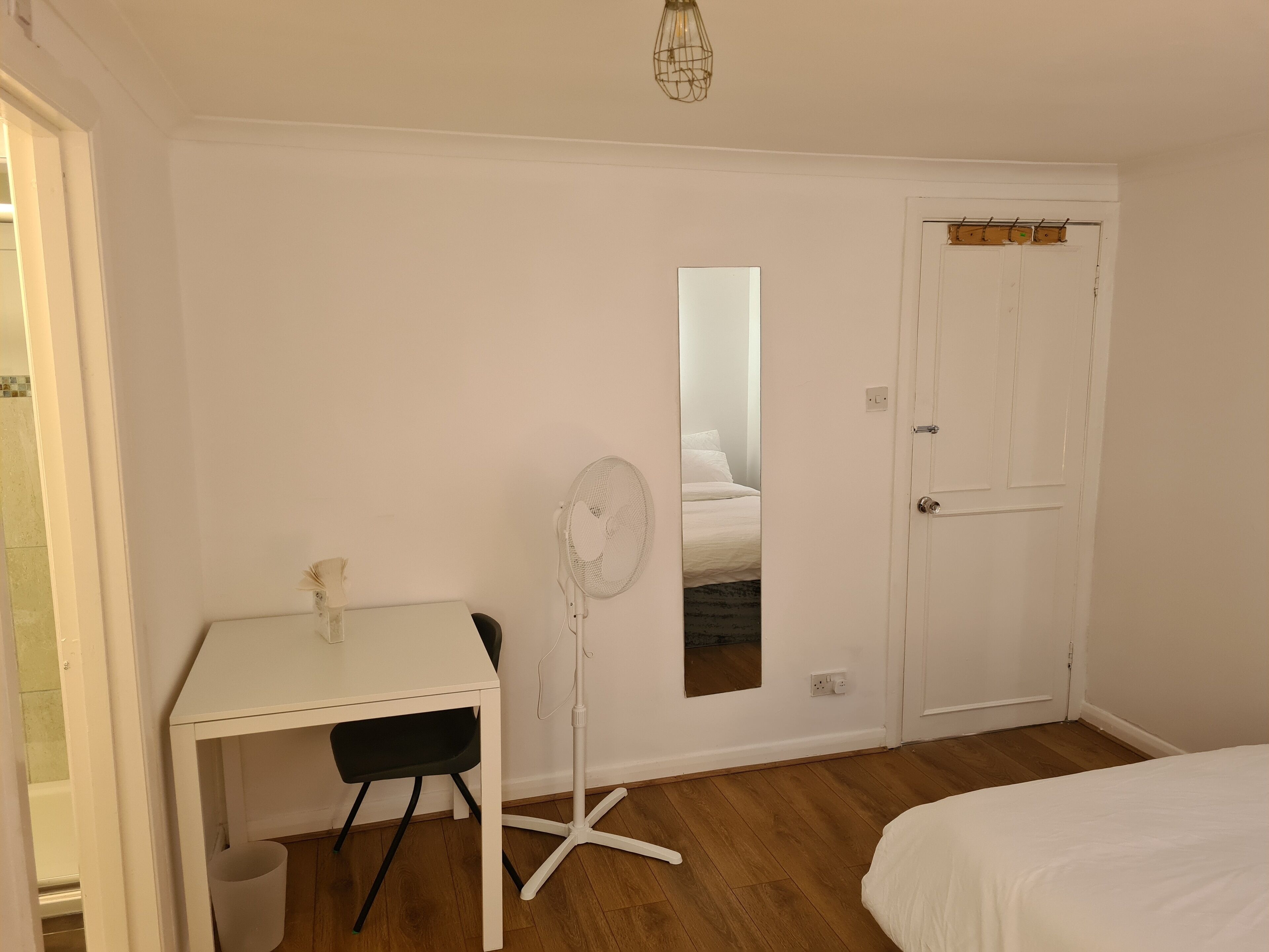 A bedroom with a white desk, a standing fan, a bed, and a door with a coat rack.