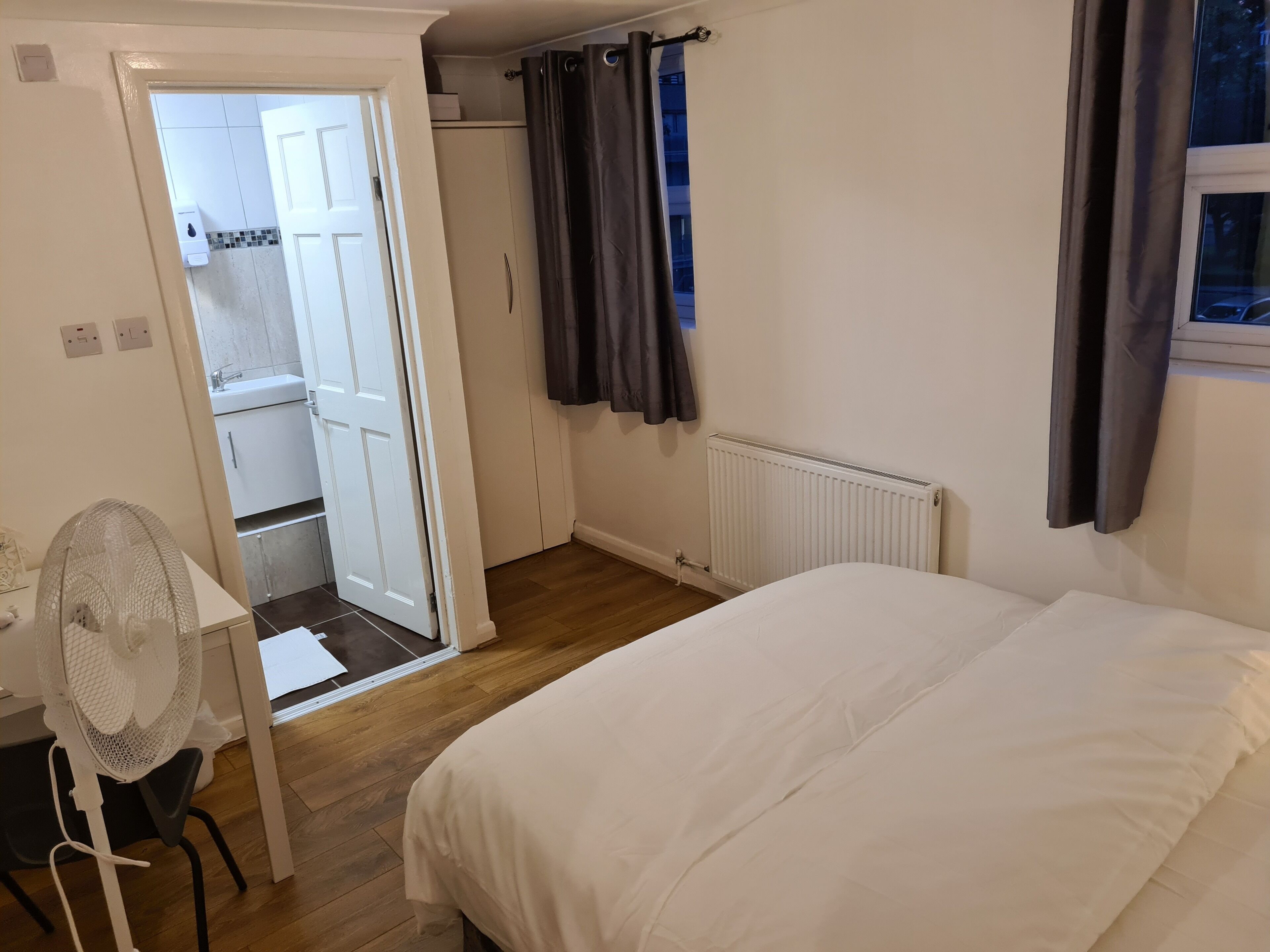 A bedroom with a bed, a desk with a chair, a radiator, and a window with curtains.