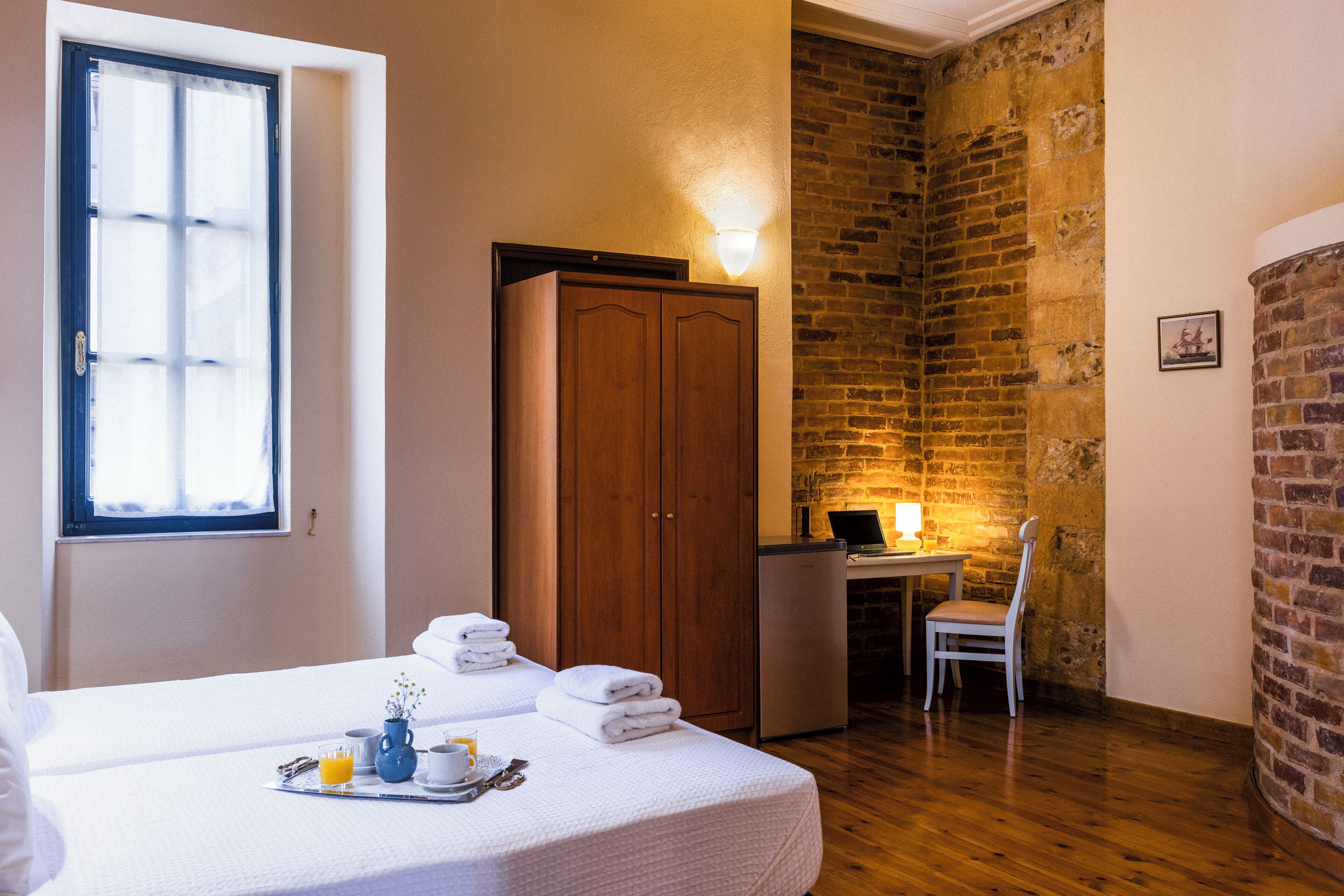 A hotel room with a bed, a desk with a computer, a chair, a small kitchenette, and a brick wall.