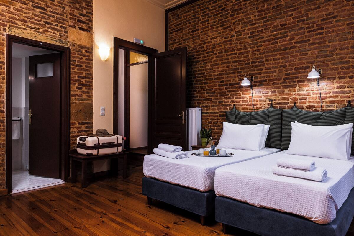 A hotel room with a brick wall, two beds with white bedding, a wooden floor, a suitcase, and a tray with a cup and a plant.
