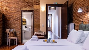 A hotel room with a brick wall, a bed with white linens, a chair, a suitcase, and a tray with two glasses of orange juice. - Sundial Lodge (Chania)