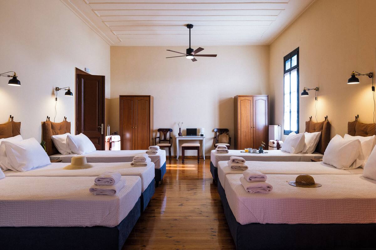 A hotel room with four beds arranged in a row, a desk, a chair, and a ceiling fan.